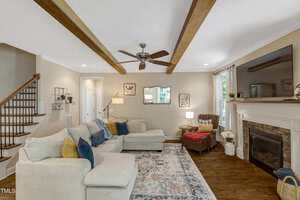 A living room with a beige sectional sofa, colorful cushions, patterned rug, stone fireplace with a TV above, armchair by the window, wooden ceiling beams, and a staircase with wooden steps and a curved railing.