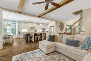 A spacious open-concept living area featuring a beige sectional sofa, patterned rug, and wooden floor. The kitchen has white cabinets, a large island with barstools, pendant lights, and a dining area by the window.