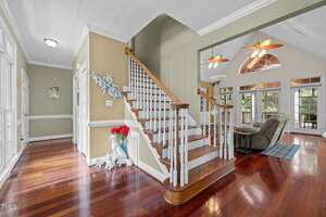 A hallway with polished wood floors and beige walls leads to a staircase with white spindles and wooden railing. To the right is a living room with armchairs, ceiling fans, and large windows letting in natural light.