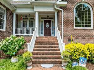 A brick house entrance with a brown door, white columns, and a set of brick steps. The porch has hanging lights and plants, with shrubs and yellow bushes on either side of the stairs. Signs are visible near the steps.