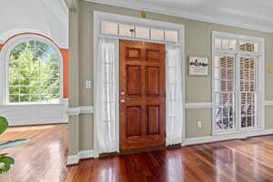 A wooden front door with glass sidelights and white curtains opens into a bright entryway with light green walls, crown molding, and hardwood floors. A large arched window and a "welcome" sign are visible.