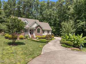 A brick house with white trim sits on a landscaped lawn surrounded by trees. A curved driveway leads to the front porch with steps and railings. Bushes and plants border the driveway and house.