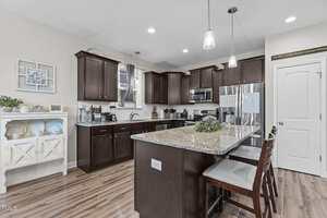 Modern kitchen with dark wood cabinets, stainless steel appliances, and a granite island with three bar stools. White walls, wood floors, and decorative shelves are also visible. Ceiling lights hang above the island.