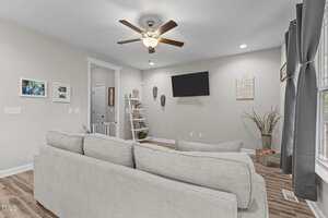 A living room with a gray sofa facing a wall-mounted TV. The room has wood flooring, a ceiling fan with lights, a small ladder shelf, framed photos on the wall, and large windows with gray curtains.