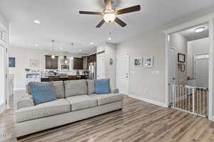 A spacious, open-concept living area with wood flooring, a gray sofa with blue patterned pillows, a ceiling fan, and a view of the kitchen with dark cabinets and pendant lights in the background.