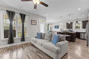 A modern living room with wood flooring, a gray sofa with blue pillows, ceiling fan, and large windows. The space opens into a kitchen with dark cabinets, a dining area, and pendant lighting.