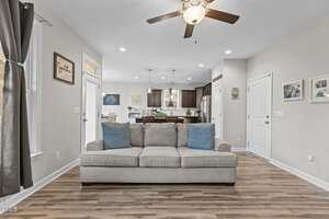 A living room with a light gray sofa featuring two blue patterned pillows, wood flooring, neutral walls, and a ceiling fan. The kitchen with dark cabinets and a dining area are visible in the background.