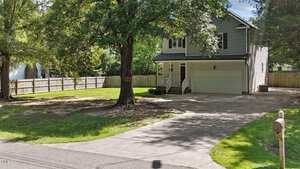 Two-story house with gray siding, an attached two-car garage, a covered front porch, and a large tree in the front yard. Concrete driveway leads to the street. Yard is bordered by a wooden fence.