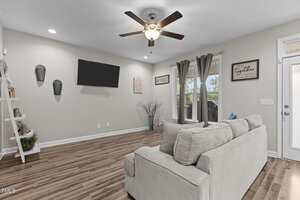A living room with light gray walls, wood flooring, a ceiling fan with lights, a wall-mounted TV, and a gray sofa. Wall art, shelves, and two windows with gray curtains are visible.