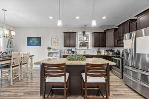 A modern kitchen with dark wood cabinets, granite countertops, stainless steel appliances, a central island with two chairs, pendant lighting, and a dining table set for four in the adjacent dining area.