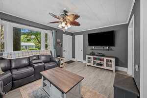 A living room with gray walls, wood-style flooring, a black leather sofa, a ceiling fan with leaf-shaped blades, a wall-mounted TV, and a window with a view of greenery outside.