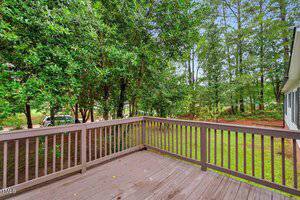 A wooden deck with a railing overlooks a grassy yard bordered by tall trees. The deck appears weathered, and the surrounding area is lush with greenery. Part of a house is visible on the right side.