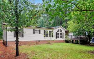 A single-story white house with black shutters and a brick foundation sits amid green trees and a grassy lawn at 38 Summerfield Lane in Henderson. There is a large front window and a wooden deck to the right side of the house.