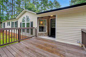 A beige manufactured home with black shutters, a large arched window, and a wooden porch deck. The front door is open, revealing a stone entryway. Tall pine trees are visible in the background.