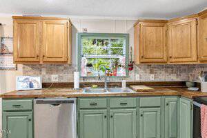 A kitchen with wood upper cabinets, green lower cabinets, a double sink under a window with plants, a dishwasher, countertop items, and a beige tile backsplash.