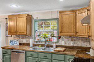 A kitchen with wood cabinets, green lower cabinets, a double sink under a window, a tiled backsplash, and a stainless steel dishwasher. Small plants and household items sit on the windowsill and counter.
