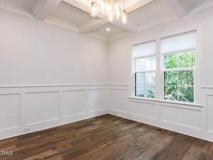 An empty room with white paneled walls, a coffered ceiling with a light fixture, two large windows with white blinds, and medium-toned hardwood flooring. Trees are visible outside the windows.