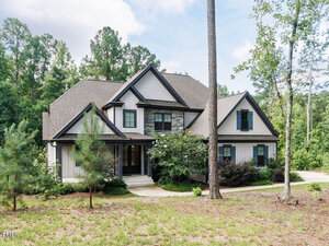 Two-story suburban house with gray siding, stone accents, black window shutters, and gabled roofs, surrounded by trees and a partly grassy, partly wooded yard under a partly cloudy sky.