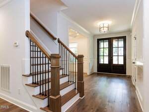 A modern home entryway featuring a wooden staircase with black spindles, hardwood floors, white walls, and double front doors with glass panels. A ceiling light fixture hangs above the foyer.