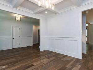 A room with light-colored walls, white wainscoting, coffered ceiling, wooden floors, and modern light fixtures. There is a doorway leading to another room and two closed doors on the left.