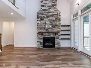 A living room with light wood floors, a floor-to-ceiling stone fireplace, built-in wooden shelves on one side, white walls, large windows, and a partial view of an upstairs railing.