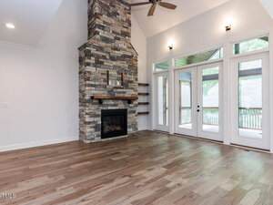 A modern living room with light wood floors, a tall stone fireplace with a wooden mantel, built-in shelves, large windows, and a glass door leading to a covered patio. Ceiling fan and wall lights are visible.