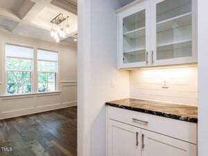 A built-in white cabinet with glass-front upper doors and a dark countertop sits under a lit backsplash; adjacent is a dining area with large windows, wood floors, and a coffered ceiling with chandeliers.