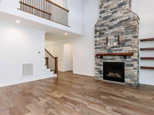 A spacious living room with light hardwood floors, a large stone fireplace with a wooden mantel, white walls, built-in shelves, and an open staircase with a wooden railing leading to an upper level.