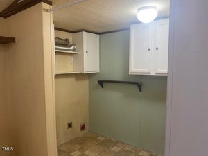 A small laundry room with light-colored walls, tile flooring, white cabinets, a wall shelf, exposed ventilation duct, and electrical outlets for laundry appliances. A ceiling light fixture is visible.