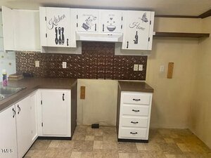 A kitchen with white cabinets, brown countertops, a metallic brown backsplash, and beige walls. Wall art on the cabinets reads "Kitchen the heart of the Home." There is an empty area between the cabinets.