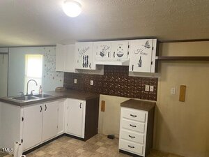 A small kitchen with white cabinets, brown countertops, a stainless steel sink, and decorative decals on the upper cabinets. The backsplash is metallic brown, and there is a ceiling light above.