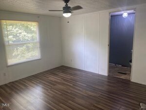 An empty room with wood-look vinyl flooring, white walls, a ceiling fan with a light, a window with blinds on the left, and a doorway leading to a bathroom with gray walls.