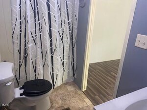 A bathroom with a white toilet featuring a black seat, a shower curtain with a black and white leaf pattern, beige tile floor, blue-gray walls, and an open door leading to a room with wood flooring.