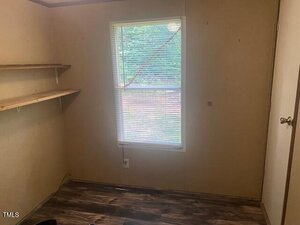 Small room with beige walls, a window with blinds, wooden shelves on one wall, and a wooden floor. The room appears empty except for the shelving and an electrical outlet near the window.