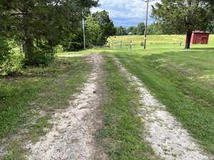 A dirt and gravel driveway bordered by grass leads toward a closed metal gate and a green field, with trees on the left and right, and a small red shed visible in the distance.