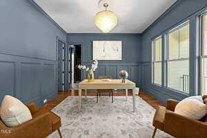 A home office with blue paneled walls, a light wood desk, a globe, a book, and a white orchid in a vase. Two brown armchairs with light pillows and a modern chandelier are also visible. Large windows line one wall.