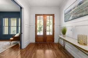 A bright entryway with double wooden front doors, white paneled walls, and a wooden floor. A narrow console table with decor sits to the right, and an adjacent room with blue walls and a brown chair is visible on the left.