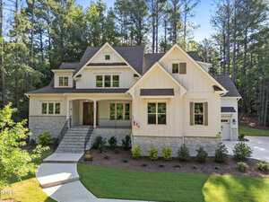 A large, two-story house with cream-colored siding and stone accents is surrounded by trees. The front entrance has steps, a covered porch, and a small American flag. A driveway curves around landscaped bushes and lawn.