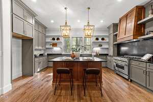 Modern kitchen with hardwood floors, gray cabinets, a wood-accented range hood, a central island with two chairs, two pendant lights, open shelves, and a window above the sink letting in natural light.