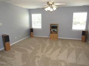 A carpeted room with light gray walls, two windows with blinds, a ceiling fan with lights, two speakers in opposite corners, and a wooden media stand with audio equipment against the far wall.