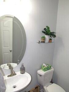 A small bathroom with a white sink, oval mirror, silver faucet, a soap dispenser, a toilet with a tissue box on top, and a glass shelf above holding two potted plants. The walls are light gray.