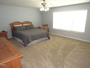 A spacious bedroom with light-colored carpet, a wooden bed with gray bedding, matching wooden side tables, a wooden rocking chair, a dresser, ceiling fan with lights, and a large window with blinds.