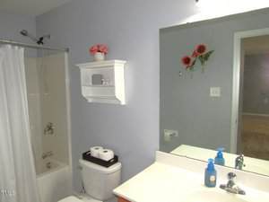 A bathroom with a bathtub and shower, a white toilet with a tray holding toilet paper, a white wall shelf with pink flowers, a sink with blue and clear soap dispensers, and a mirror reflecting red flowers on the wall.