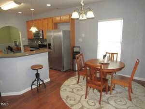 A kitchen and dining area with wood cabinets, stainless steel appliances, a round wooden table with four chairs on a patterned rug, and a small bar with a stool; pendant and ceiling lights add illumination.