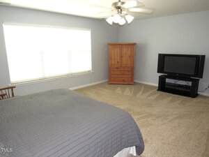 A bedroom with light gray walls, beige carpet, a bed with a gray comforter, a wooden armoire, a flat-screen TV on a stand, a wooden chair, and a ceiling fan with lights. Sunlight comes through a large window.