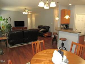 Open-plan living area with wood flooring, a round dining table in the foreground, a kitchen with a breakfast bar and stools to the right, and a living room with dark sofas and a TV in the background.