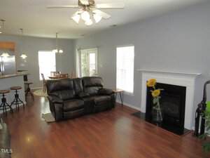 A living room with dark leather sofas, hardwood floors, a white fireplace with a vase of sunflowers, and a ceiling fan. The space opens to a kitchen with barstools and a dining area with windows and glass doors.