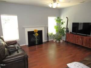 A living room with wood flooring, brown leather sofas, a black fireplace with a sunflower vase, a ceiling fan with lights, two windows, several potted plants, and a flat-screen TV on a wooden stand.