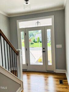 A bright entryway with a glass front door and side windows, hardwood floors, gray walls, white trim, a staircase with dark handrails, and a view of the street and greenery outside.