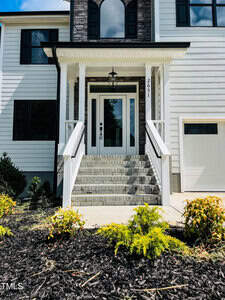 Front view of a house with a white exterior, stone steps leading up to a glass front door, two railings, black-trimmed windows, a small garden with shrubs, and part of a garage door visible on the right.